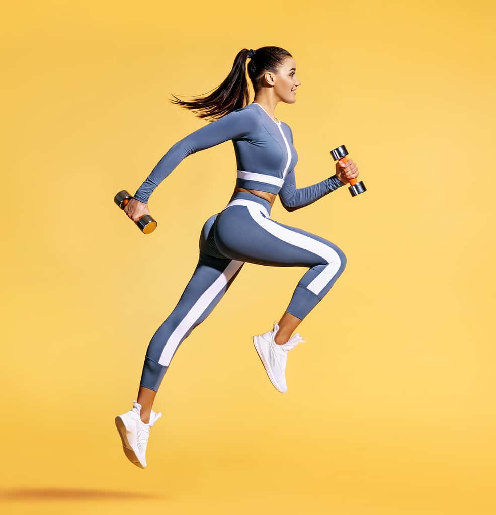 Sporty woman jumping with dumbbells. Photo of active woman in sportswear on yellow background. Dynamic movement. Side view. Sport and healthy lifestyle